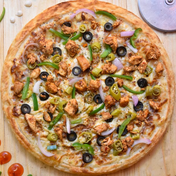 A whole chicken pizza with olives, green peppers, onions, and chili flakes on a wooden table.